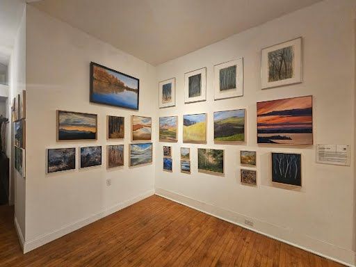 Corner of the gallery with framed landscape paintings hung salon-style